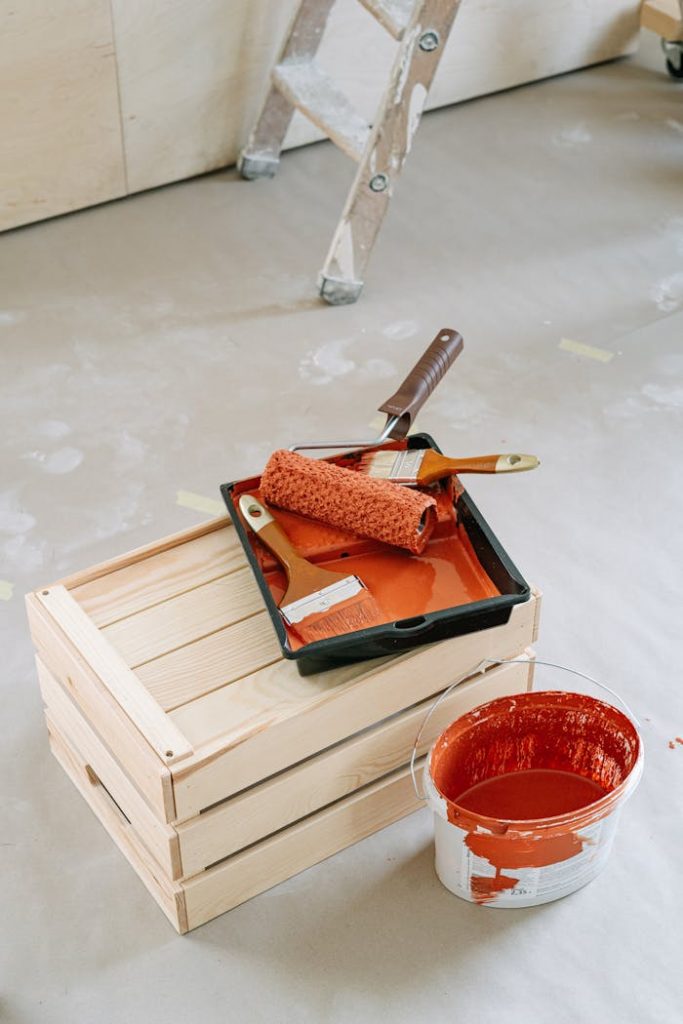 A close-up of home renovation tools including paint roller and red paint bucket on a wooden crate.